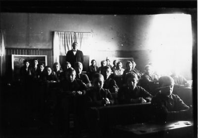 Lärare stående längst bak i skolklass med barn sittande i sina bänkar.
