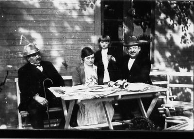 2 män och 1 kvinna sittande och 1 flicka stående vid trädgårdsmöbel.
Nyckelord: Käpp