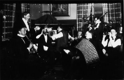 8 personer stående eller sittande med instrument i lokal.
