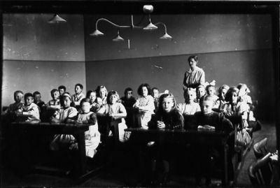 Lärarinna stående längst bak i skolklass med barn sittande i sina bänkar.
Nyckelord: Skolklass