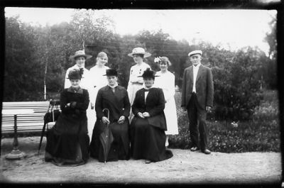 Man och 4 kvinnor stående bakom och 3 kvinnor sittande på parkbänk.
Nyckelord: Parkbänk Paraply