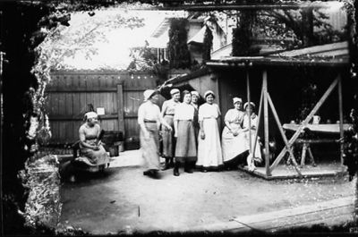 5 kvinnor stående och 3 sittande framför plank.
Nyckelord: Bakgård