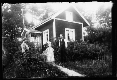 Man, kvinna och flicka stående i trädgård framför byggnad.
Nyckelord: Trädgård