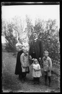 Man, kvinna, med barn i famnen och 3 barn stående i trädgård.
