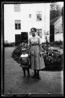 Kvinna och barn stående på gårdsplan med byggnad i bakgrunden.
