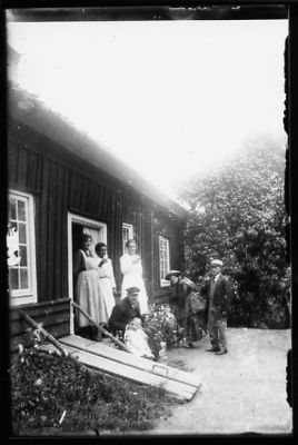 Man och 4 kvinnor stående, man och flicka sittande framför byggnad.
Nyckelord: Källarlucka