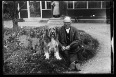 Man och hund sittande på backen framför byggnad.
Nyckelord: Hund