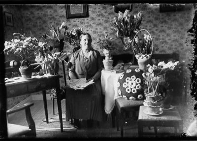 Kvinna sittande bland blommor i rum.
Nyckelord: Blommor