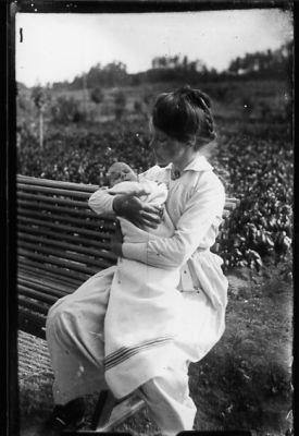 Kvinna sittande på parkbänk med baby i famnen.
Nyckelord: Parkbänk
