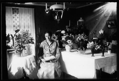 Kvinna sittande i rum bland blommor.
Nyckelord: Blommor Lampa Duk