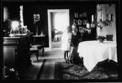 Man och kvinna sittande och flicka stående vid bord i rum.
Nyckelord: Lampa Byrå