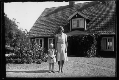 Kvinna och flicka stående på gårdsplan framför byggnad.
