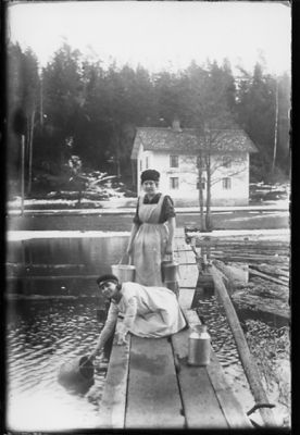 2 kvinnor hämtar vatten. 1 står på brygga och 1 står på knä på brygga.
Nyckelord: Vatten Kruka Hink