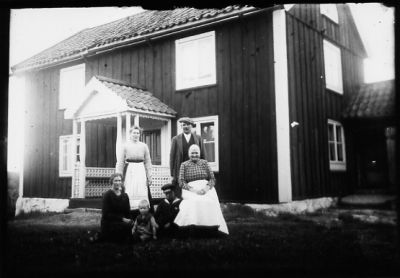 Man och kvinna stående, 2 kvinnor och 2 barn sittande framför byggnad.
Nyckelord: Bostad