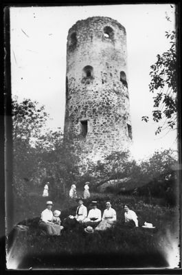 Personer sittande i gräset framför ruin.
