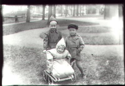 2 barn stående och 1 barn sittande i barnvagn i park.
Nyckelord: Barnvagn Park