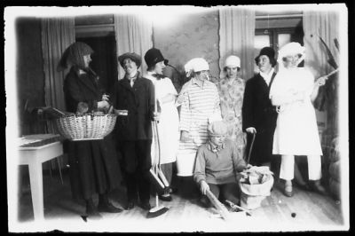 7 utklädda stående och 1 sittande kvinna i rum.
Nyckelord: Korg Käpp