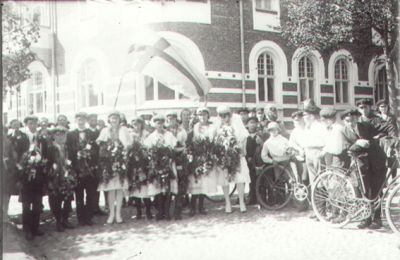 Ett antal examenselever stående på gatan framför byggnad.
Nyckelord: Blommor Cykel Skolmössor Flagga