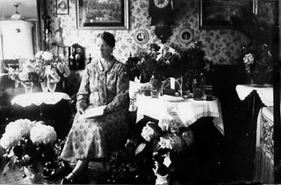 Kvinna sittande i finrum med blommor runt omkring sig.
Nyckelord: Blommor Tapet Klocka Tavla