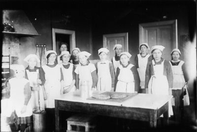 Kvinna och 12 flickor stående bakom bord.
Nyckelord: Dräkt Spisel Bröd Mjölkkruka