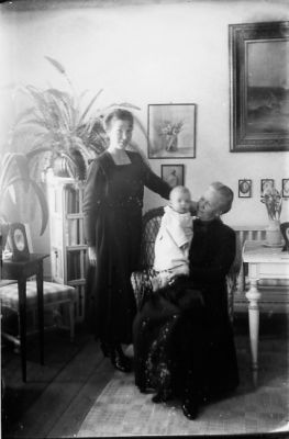 Kvinna stående och kvinna sittande med barn i famnen, i finrum.
Nyckelord: Tavla Pedistal