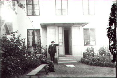 Man stående vid trappa och kvinna sittande på bänk framför byggnad.
Nyckelord: Trädgård Bänk