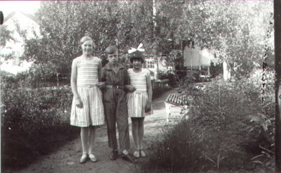 Pojke och 2 flickor stående på gång i trädgård.
Nyckelord: Trädgård Parkbänk Bostad