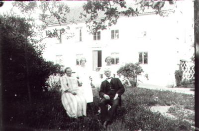 Man och kvinna sittande, 2 kvinnor stående i trädgård framför byggnad.
Nyckelord: Trädgård