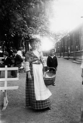 Kvinna stående med korg på gårdsplan.
Nyckelord: Parkbänk Korg Dräkt