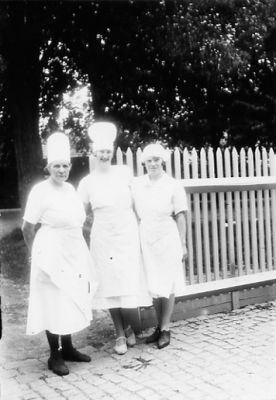 3 kvinnor stående vid staket.
Nyckelord: Staket Gatsten Kockmössa