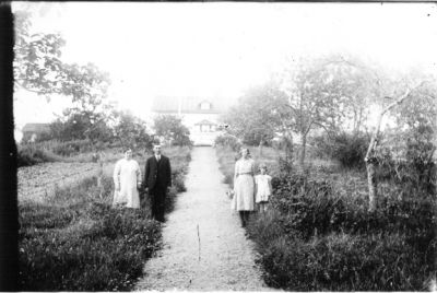 Man, 2 kvinnor och 1 flicka stående vid grusgång i trädgård med byggnad i bakgrunden.
Nyckelord: Gång Byggnad