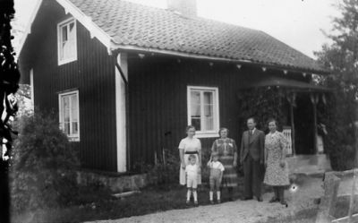Man, 3 kvinnor och 2 barn stående framför byggnad.
