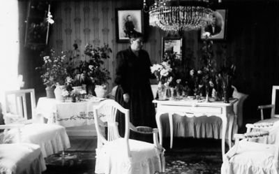 Kvinna stående i finrum bland blommor.
Nyckelord: Högtid Blommor Lampa Tavla