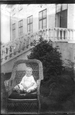 Barn sittande i korgstol framför byggnad.
Nyckelord: Korgstol Trappräcke