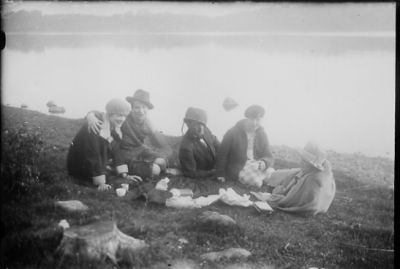 Man och 4 kvinnor sittande intill vatten.
Nyckelord: Picnic