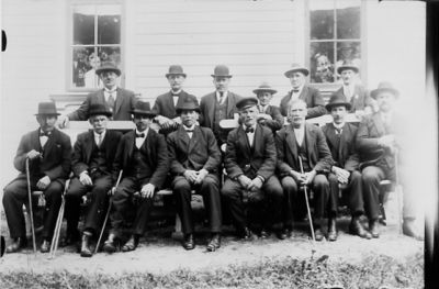 8 män sittande på bänk och 6 män stående bakom.
Nyckelord: Käpp Bänk Husvägg Byggnad