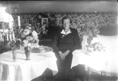 Kvinna sittande vid bord i rum.
Nyckelord: Väggbonad Blommor