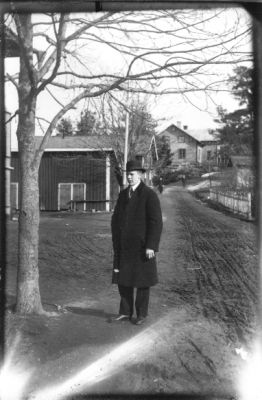 Man stående vid träd med byggnader i bakgrunden.
Nyckelord: Väg Byggnad