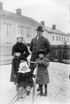 Man, kvinna, pojke och flicka stående vid spark med flicka sittande på.
Nyckelord: Spark Staket Bostad Vinter