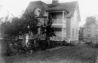 2 män, 1 kvinna och 3 barn stående i trädgård framför byggnad.
Nyckelord: Trädgård Balkong