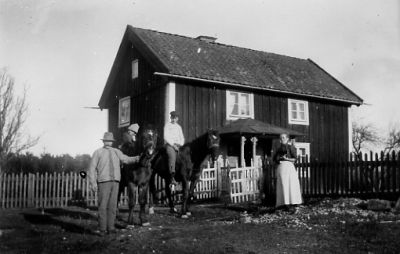 Man och kvinna stående och 2 pojkar sittande på varsin häst, framför tomt.
Nyckelord: Häst Staket