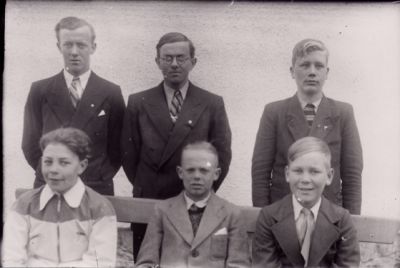 3 män stående bakom och 3 pojkar sittande på bänk.
Nyckelord: Bänk Husvägg