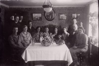 3 män och kvinna stående och 3 män och 5 kvinnor sittande vid bord, i rum.
Nyckelord: Blommor