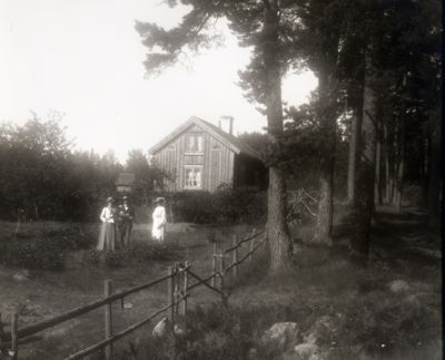 2 kvinnor och 1 man stående i hage intill torpstuga
