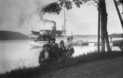 Familj sittande vid strandkant med båt liggande vid brygga.
