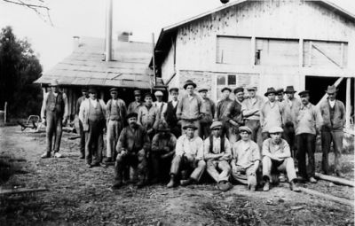 Ett antal män stående och sittande framför byggnad.
