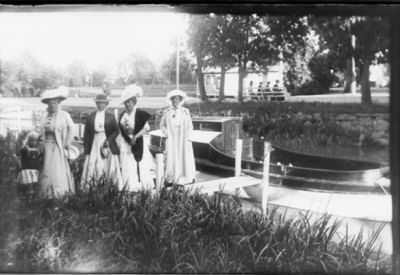 4 kvinnor stående vid vattendrag.
Nyckelord: Brygga Dräkt Hatt