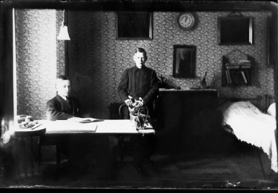 Pojke sittande och pojke stående bakom skrivbord.
Nyckelord: Skrivbord Tapeter Klocka
