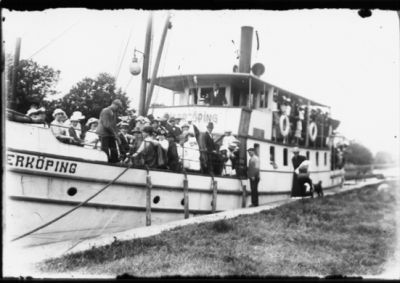 Ett antal personer stående på båt och på kaj vid sluss.
