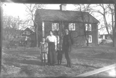 Man, kvinna och 2 barn stående i trädgård framför byggnad.
